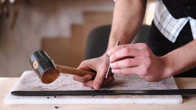 A leather craftsman is making holes in a hand-made belt. The tanner is using a mallet and a hole punch.