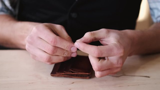 A tanner is stitching a hand-made leather wallet. The currier is using a needle with thread.