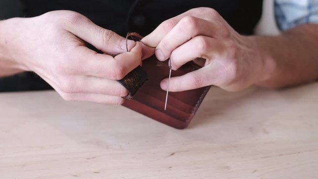 A tanner is stitching a hand-made leather wallet. The currier is using a needle with thread.