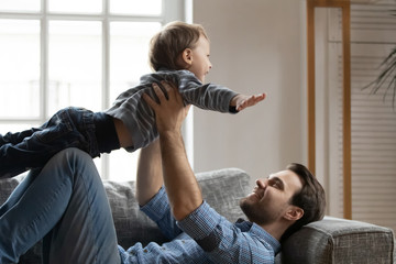 Smiling strong dad lying on comfortable sofa, lifting small preschool son. Excited little kid boy...