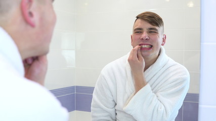 A young man in a white coat looks in the mirror at his teeth. View through the mirror