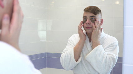 A young man puts cosmetic patches on his face. Spa treatments. Brown hydrogel patches for skin rejuvenation. He is standing in a white bathrobe in front of the bathroom mirror. View through the mirror