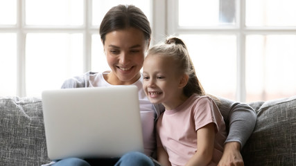 Head shot young smiling mother relaxing on comfy sofa with small school aged daughter, watching funny videos on laptop. Attractive happy nanny babysitter showing educational application to kid girl.