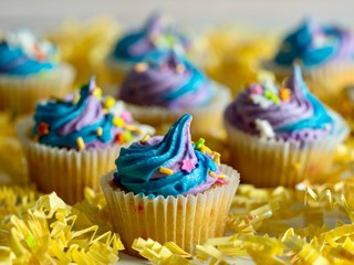 Mini yellow cupcake with blue and purple swirly frosting decorated with colorful confetti sitting on a bed of yellow paper shreds.  Festive and delicious for a celebration.