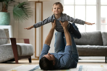 Full length young strong dad lying on floor carpet, lifting small playful child son in air. Smiling little kid boy imagining plane, making exercise on trust with father in modern living room.