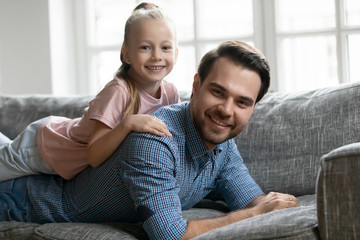 Fototapeta na wymiar Portrait of smiling pleasant young father lying on comfy sofa, holding cute little blonde kid daughter on back. Affectionate two generations family enjoying weekend time, resting on couch at home.