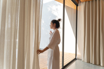 Fototapeta na wymiar Asian woman in bathrobe suit open curtain to see view outside in morning, Travel and Holiday concept