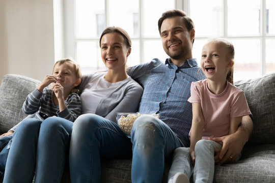 Happy young married couple embracing little kids siblings, watching funny movies or tv show, eating popcorn, resting on sofa. Smiling family enjoying weekend time with small kids in living room.