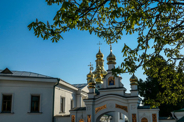 A close-up view on Pechersk Lavra in Kiev, Ukraine, known as the Kiev Monastery of the Caves. Trees blocking the view. Green rooftops with golden domes. Walls are painted white.