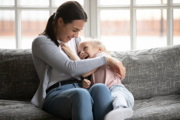 Happy young mother tickling little blonde daughter, having fun on comfortable sofa at home. Overjoyed small child girl playing with laughing nanny babysitter elder sister, enjoyin weekend time.