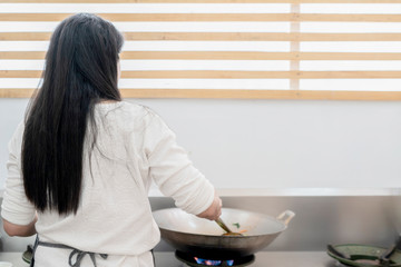 Asian female with long black hair cooks food in steel pan on stove with copy space behind.