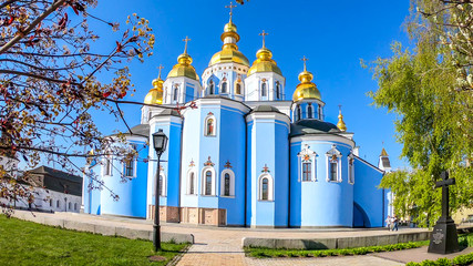 Fototapeta na wymiar St Michael's Golden-Domed Cathedral in Kiev, Ukraine, seen from the back, park side. The walls of the cathedral are painted blue and nicely decorated on each facade. Golden domes reflecting the sun