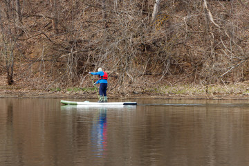 A man in a red vest is rowing while standing on a board (SUP) along a river.