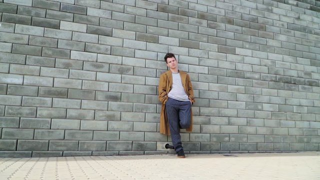 Beautiful young fashionable guy standing beside the concrete wall in the city Park. On it wearing a long brown coat. Slow motion. Shooting on the Steadicam
