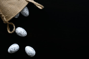 Top view of easter decoration. White eggs with painted silver details, scattered from jute bag. Black background. Copy space.