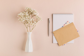 Twigs of gypsophila in a white vase, notebook, pen and envelope on a beige pastel background. Greeting card concept. Top view