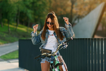 Attractive tempting young girl in fashion jeans clothes wears stylish glasses ,sitting on the bicycle and posing on camera at summer evening. Slow motion