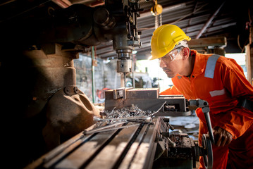 Workers in the shop wear orange work clothes and wear glasses to prevent metal chips from penetrating. Stand beside the drilling rig and use metal drills in industrial plants - obrazy, fototapety, plakaty