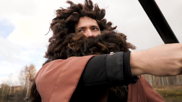 Close up of a man with fake beard and a wig sitting in the boat and rowing with a paddle on trees and cloudy sky background. Stock footage. Young guy looking like an old man while rowing with an oar.