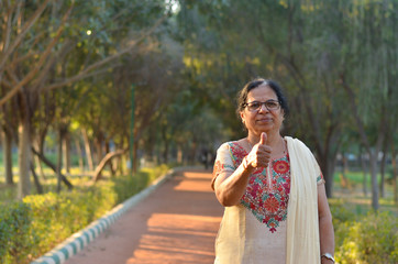 Fototapeta premium Portrait shot of a happy looking senior north Indian woman wearing traditional chikan kari Indian salwar kameez showing a thumbs up in a garden against a bokeh of canopy of trees.