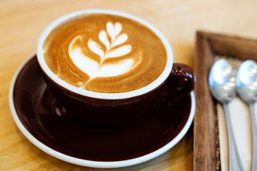 Hot latte coffee with latte art in black cup on wood table background next to spoon set in café restaurant, beautiful steamed milk by overhead angle pouring.