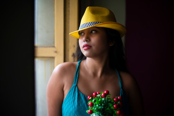 Portrait of an Asian/Japanese/Korean brunette young girl in greenish blue  western dress and yellow hat holding cherry tree front of a window inside of a room. Fashion and cosplay photography.
