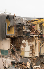 Demolishing a building with a large backhoe. Overcast day. Air is dusty. Lots of debris from the demolition. Debris falls as building is being demolished.
