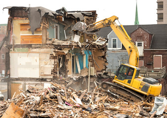 Fototapeta premium Demolishing a building with a large backhoe. Overcast day. Air is dusty. Backhoe is on debris from the demolition. Debris falls from a wall being demolished.