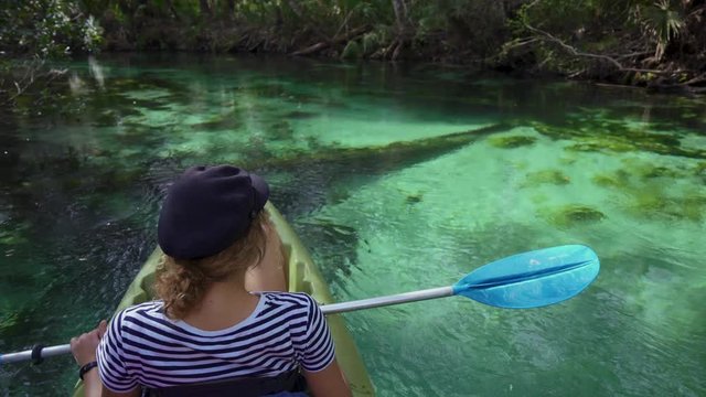 A young brunette model girl with curly hair is paddling a kayak through the magical clear blue water Weeki Wachee State Park spring river. The sun is shining through jungle trees during vacation.