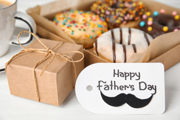 Tag with phrase HAPPY FATHER'S DAY, donuts, coffee and gift on white wooden table