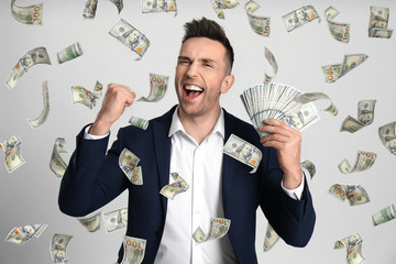 Happy young man with dollars under money rain on light background