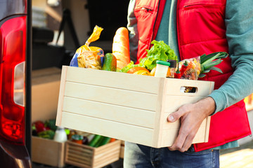 Courier holding crate with products near car outdoors, closeup. Food delivery service