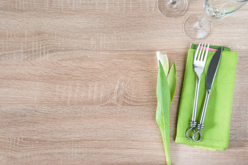 Table setting with white tulip, forged fork and knife on green napkin, set of wineglasses on wooden background. Space for text, top view