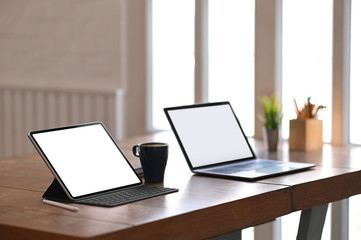 Photo of white blank screen computer putting on wooden table together with potted plant, coffee cup, coffee cup, white blank screen computer tablet. Vintage style workplace concept.