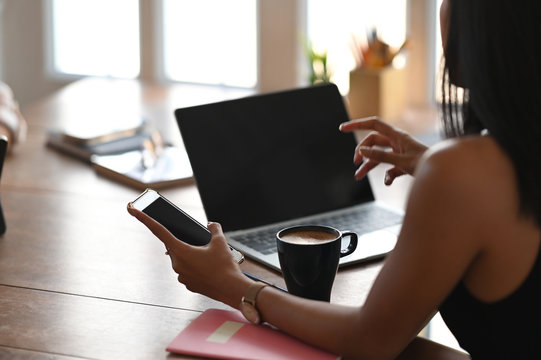 Cropped image of beautiful woman holding a black blank screen smartphone in hand while sitting at the wooden working table with living room windows as background.