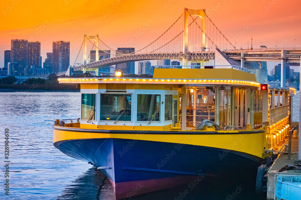 Wall mural Japan: Tourist boat in Tokyo. Rainbow Bridge in Japan. Passenger ferry glows on the background of the evening city. Ferry on background of Japanese skyscrapers. Traveling along the Tokyo Bay by ship