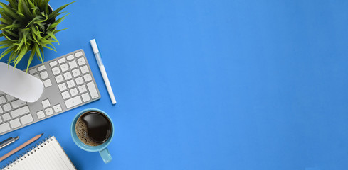 Top view Simple panorama workspace. Flat lay coffee cup, pens, pencil, notebook, keyboard, wireless mouse and potted plant on modern blue working table. Orderly workplace concept.