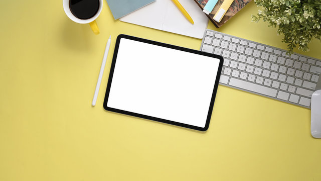 Top view image of workspace with flat lay white blank screen computer tablet, coffee cup, stylus pen, marker pens, stack of notebook, keyboard, mouse and potted plant on colorful working table.