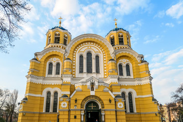 A view on the front facade of St Volodymyr's Cathedral. The cathedral is painted yellow, with blue and golden-domed rooftop. ON the side of the Cathedral there are trees. Overcast and cold day.
