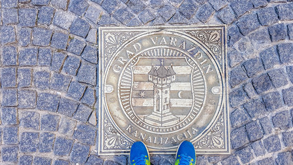 Sewage well with a city emblem of Varazdin and a sign Kanalizacja, meaning sewage. A pair of shoes standing on the well. Street paved with cobblestones.
