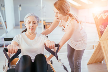 Respectable old age grey hair beautiful woman workout pilates with personal trainer on reformer practice in pilates studio, working out indoor, correcting beginners, rehabilitation concept.