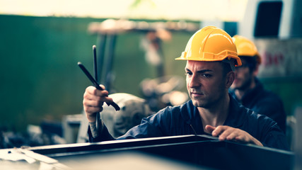 Scene of industrial workers wearing yellow safety hat while holding tool in his hand show moody curious face with blurry another worker behind in industrial atmosphere.