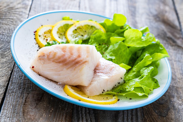 Fresh raw cod with vegetable leaves and lemon on wooden table