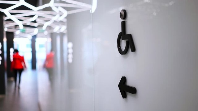 person in jacket walks along modern department store with black sign for disabled on white wall under ceiling with illumination decorations backside view