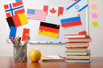 Stack of books, pencils, an apple in front of school whiteboard with flags of different countries....