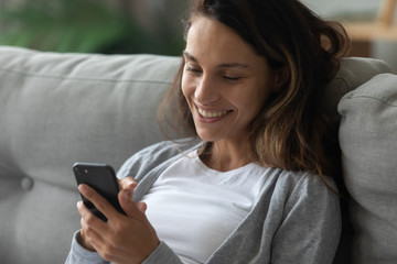 Smiling young woman rest on cozy sofa in living room using modern cellphone texting or messaging, happy millennial girl relax on comfortable couch at home browsing wireless internet on smartphone