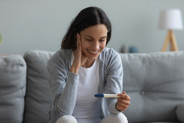 Smiling young woman sit on couch in living room look at positive pregnancy test feel excited, happy...
