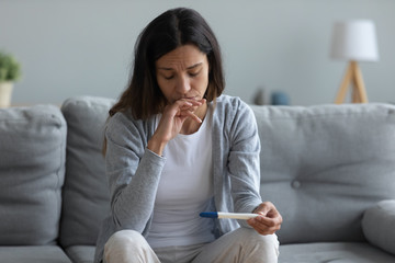 Upset millennial girl sit on couch in living room feel frustrated with bad news pregnancy test...