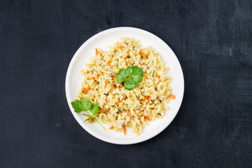 Stewed side dish of brown rice with onions and carrots and celery in a white plate on a black background. Traditional national cuisine. Copy space