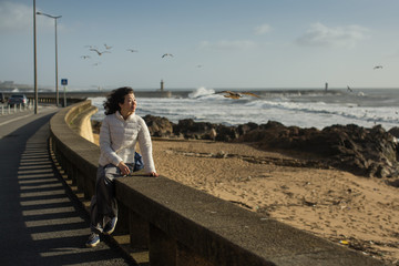 Beautiful young multicultural asian woman sitting of sea promenade. Seagulls fly over the foaming surf.